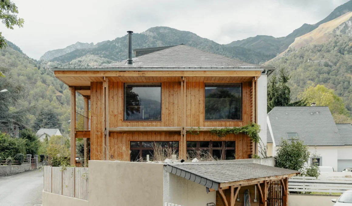 Exterior view of a two-story renovated house in the French Pyrenees, featuring vertical wood cladding, a large overhanging timber structure, and a slate mansard roof against a backdrop of mountains.