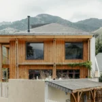 Exterior view of a two-story renovated house in the French Pyrenees, featuring vertical wood cladding, a large overhanging timber structure, and a slate mansard roof against a backdrop of mountains.