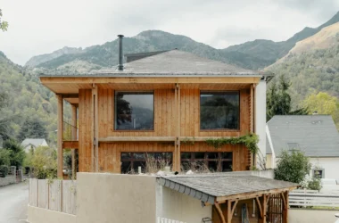Exterior view of a two-story renovated house in the French Pyrenees, featuring vertical wood cladding, a large overhanging timber structure, and a slate mansard roof against a backdrop of mountains.