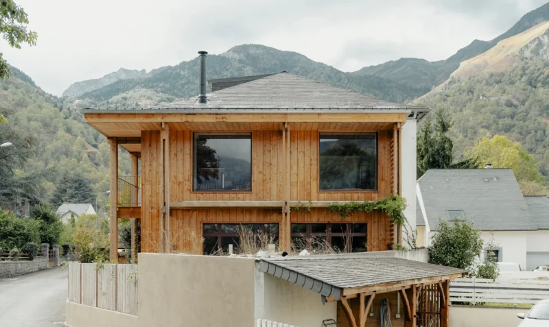Exterior view of a two-story renovated house in the French Pyrenees, featuring vertical wood cladding, a large overhanging timber structure, and a slate mansard roof against a backdrop of mountains.