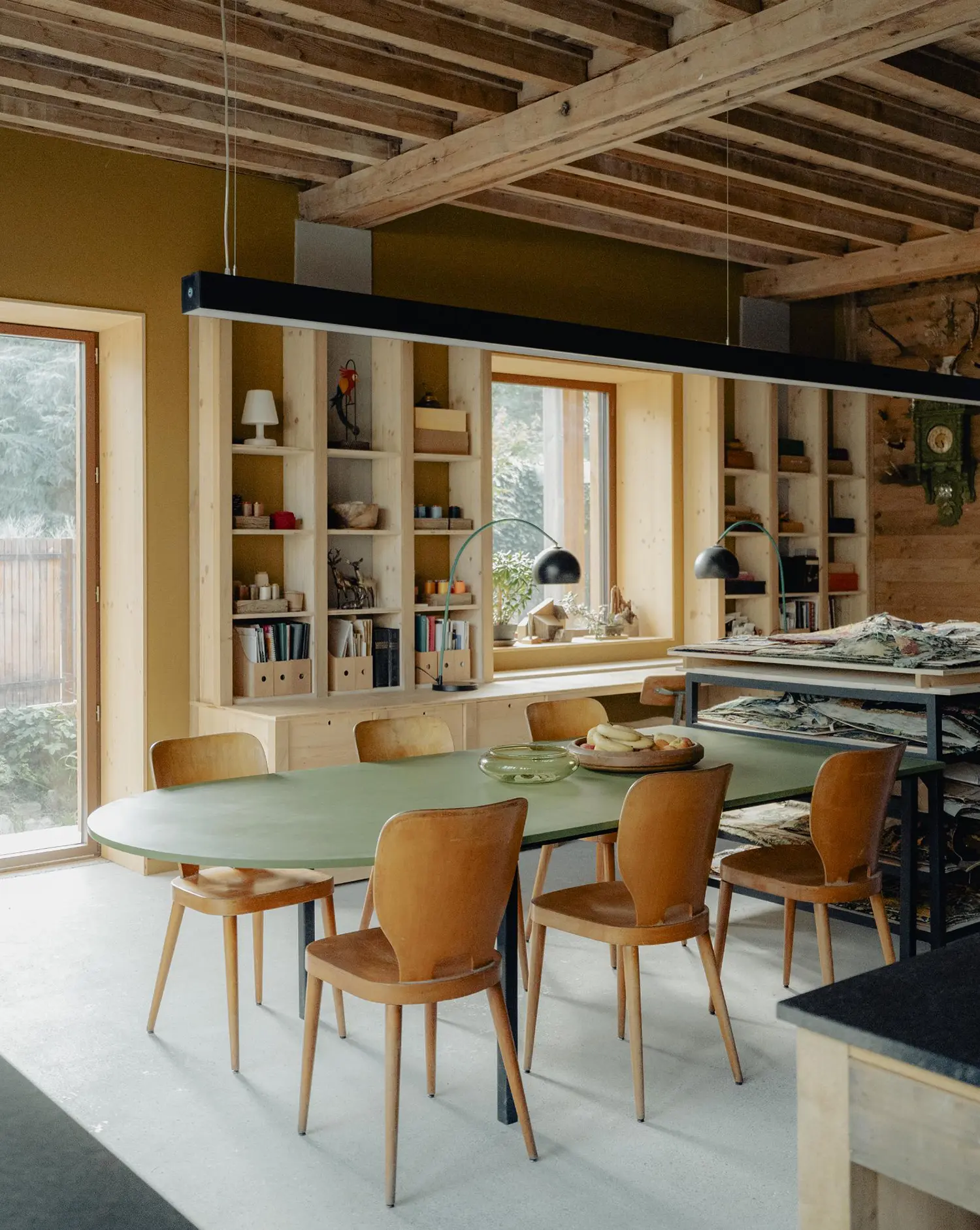 Interior view of a dining area with an oval green table and vintage wooden chairs, backed by built-in light wood shelving. The space features exposed timber beams and ochre-colored walls.