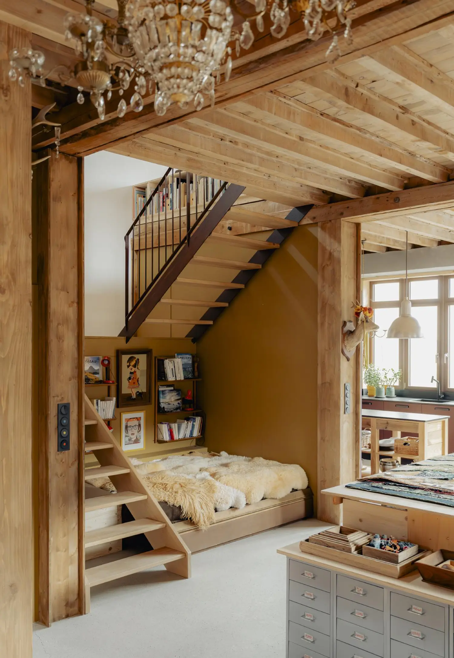 View of a central light-wood staircase with black metal balustrades ascending towards a mezzanine with integrated bookshelves. Below, a reading nook with a sheepskin-covered daybed is nestled against a mustard-yellow wall. A crystal chandelier hangs overhead.