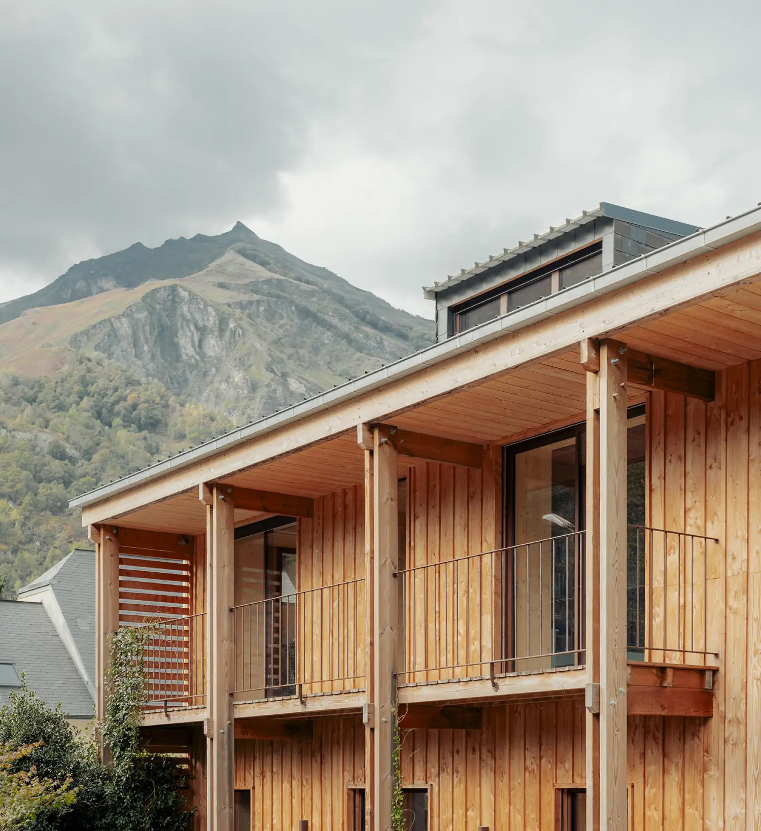 Exterior detail view of a stacked two-story timber terrace with slim metal balustrades, cantilevered under a wide, metal-clad overhang against a jagged mountain peak.