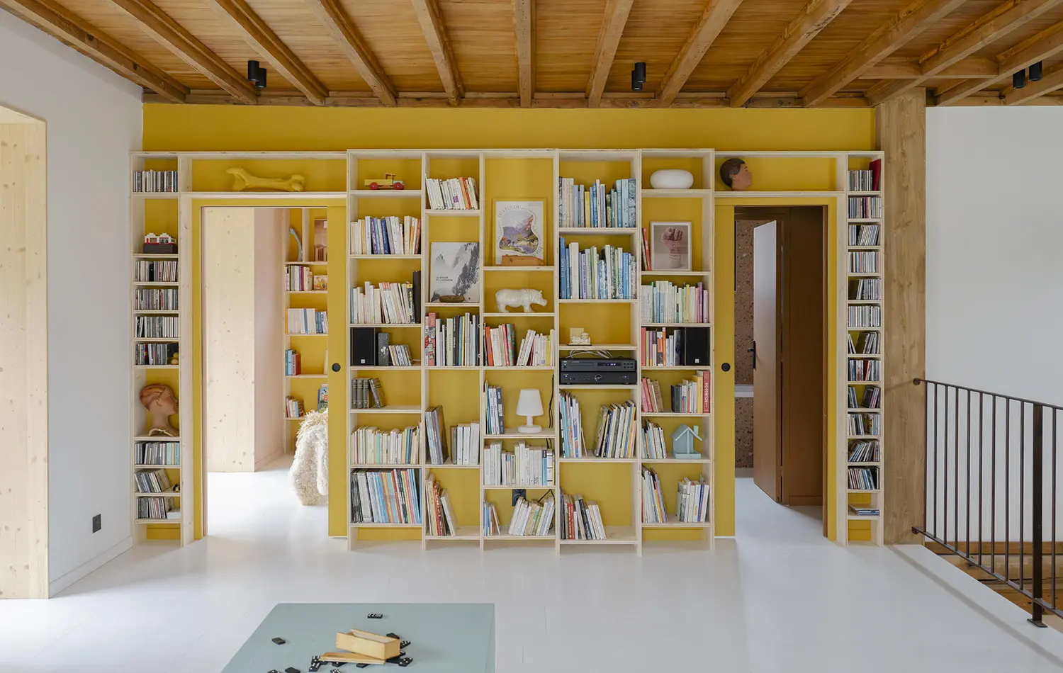 Bright upper-floor landing with a continuous, brightly painted yellow wall that integrates multiple doorways and an extensive floor-to-ceiling shelving unit filled with books, vinyl records, and personal artifacts.