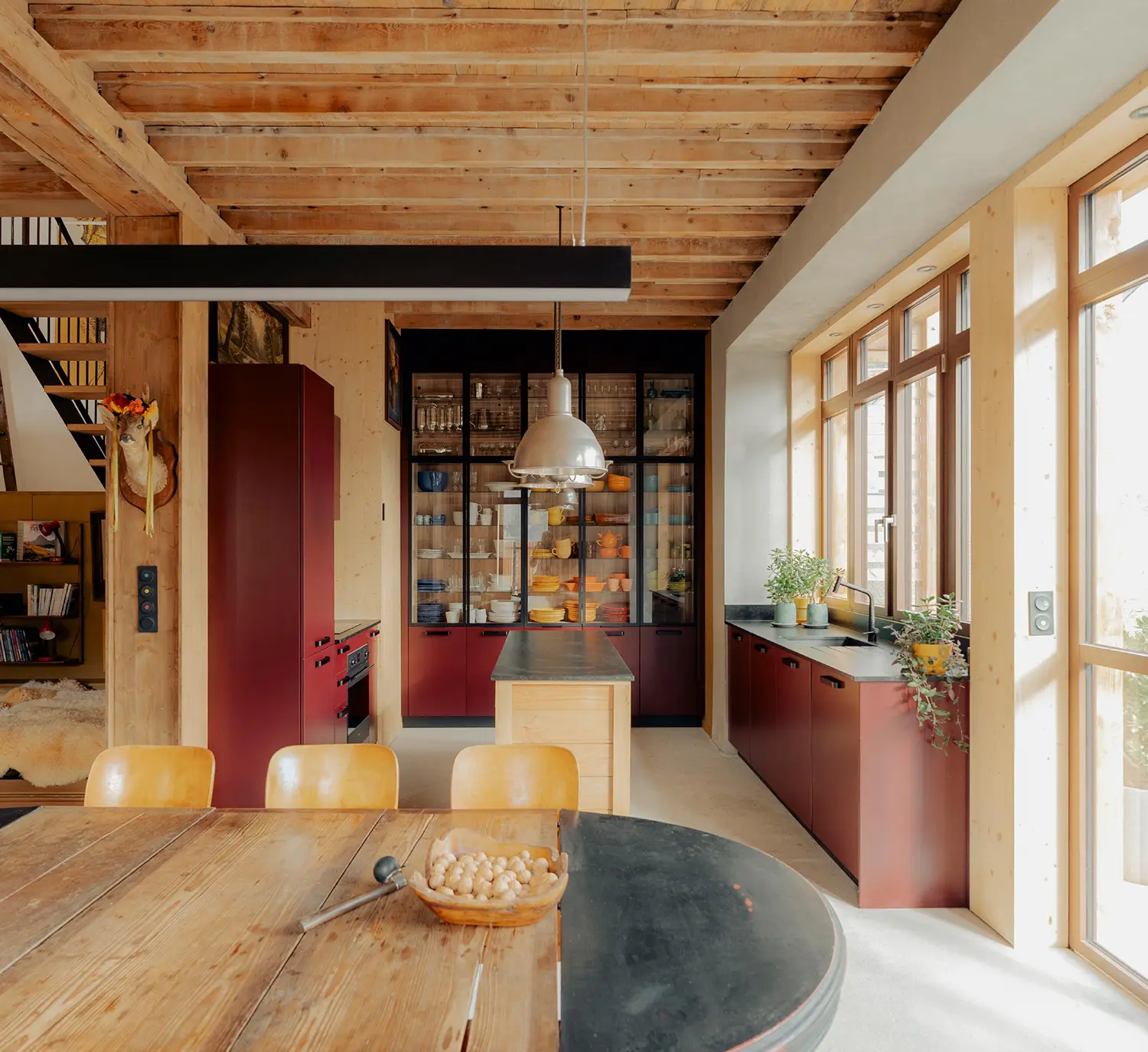 Detailed view of a kitchen with deep maroon-red cabinetry, dark slate countertops, and a central natural-wood island. The back wall features a large glass and black-frame display unit. Vintage wooden chairs are placed around a large table in the foreground.