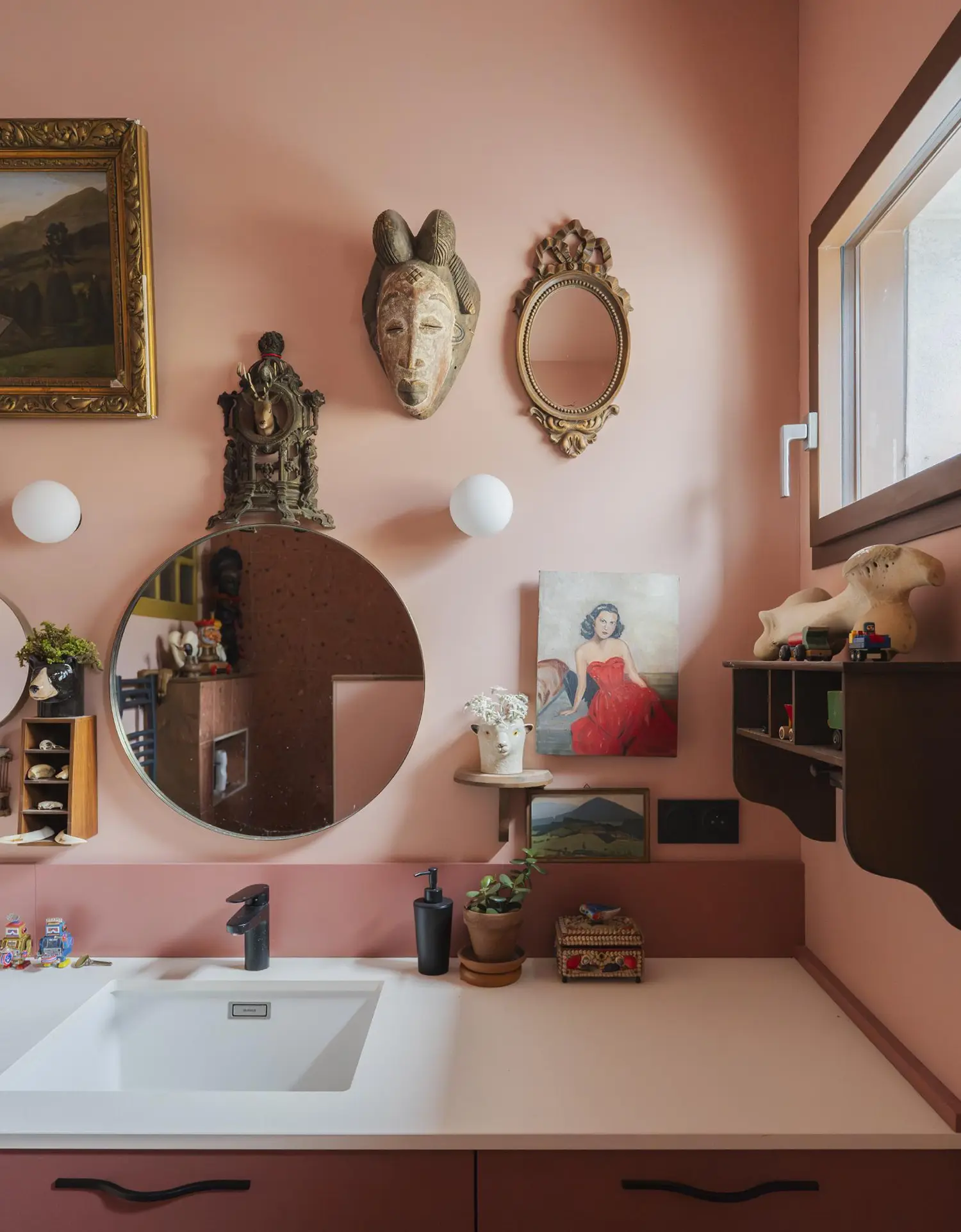 Detailed view of a bathroom vanity area painted entirely in soft dusty pink. The wall features multiple curated elements, including two mirrors (one ornate, one round), a wooden African mask, various framed paintings, and small ceramic figures.