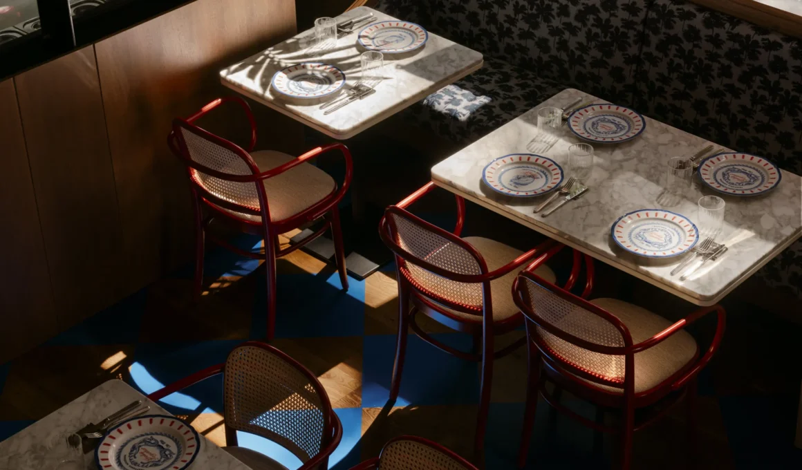 Overhead view of the Like Mike trattoria in Milan, showcasing high-contrast dappled sunlight on red Viennese straw chairs, Carrara marble tables with patterned plates, and a unique blue resin checkered floor set against dark wood.