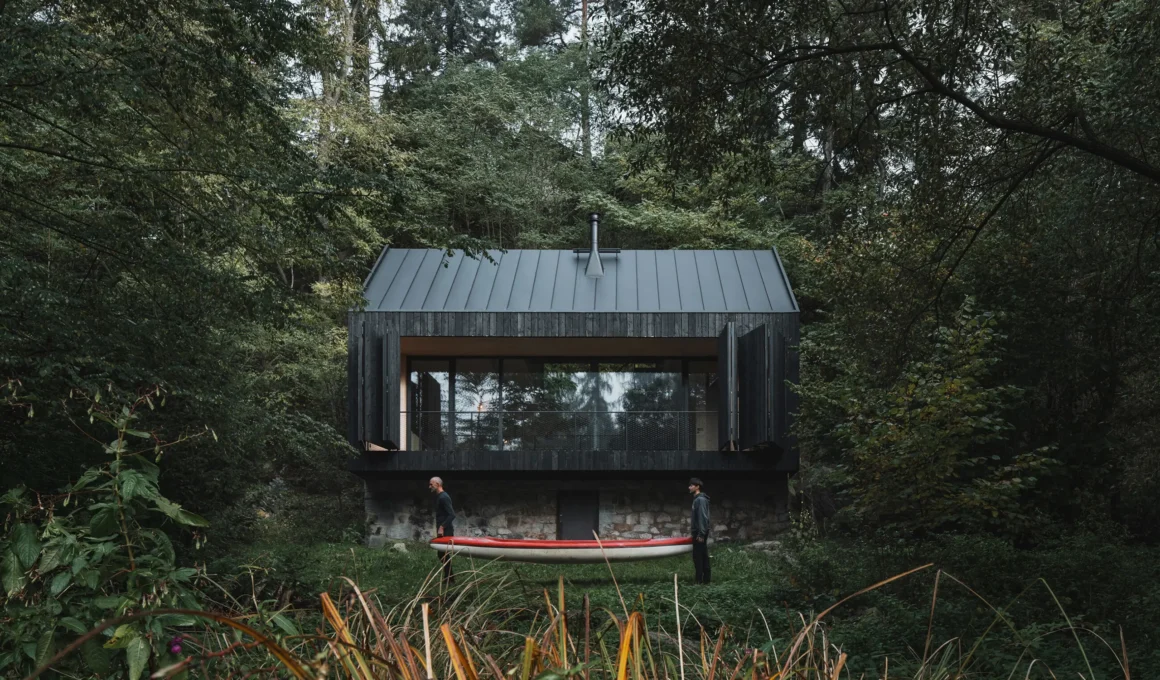 A blackened timber cabin on a stone base nestled in a lush green forest with two people carrying a red canoe in the foreground.