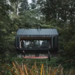 A blackened timber cabin on a stone base nestled in a lush green forest with two people carrying a red canoe in the foreground.