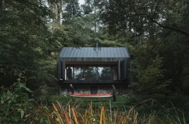 A blackened timber cabin on a stone base nestled in a lush green forest with two people carrying a red canoe in the foreground.