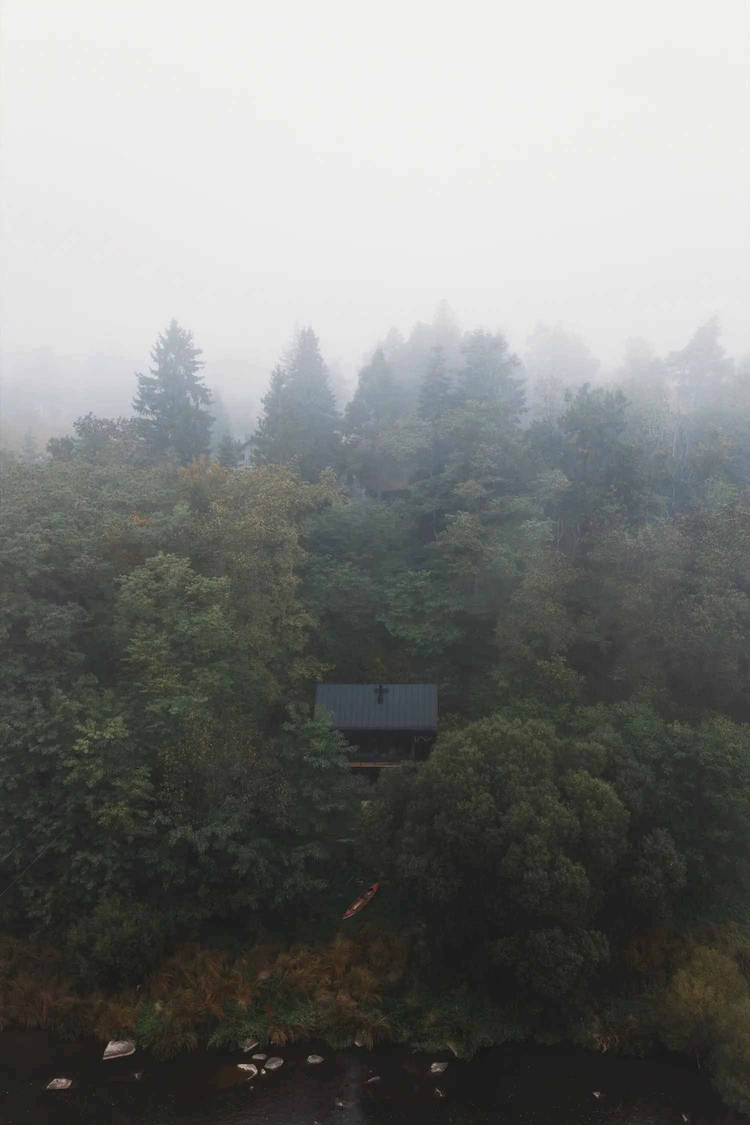 An aerial view of a dark cabin surrounded by a thick, misty forest next to a river.