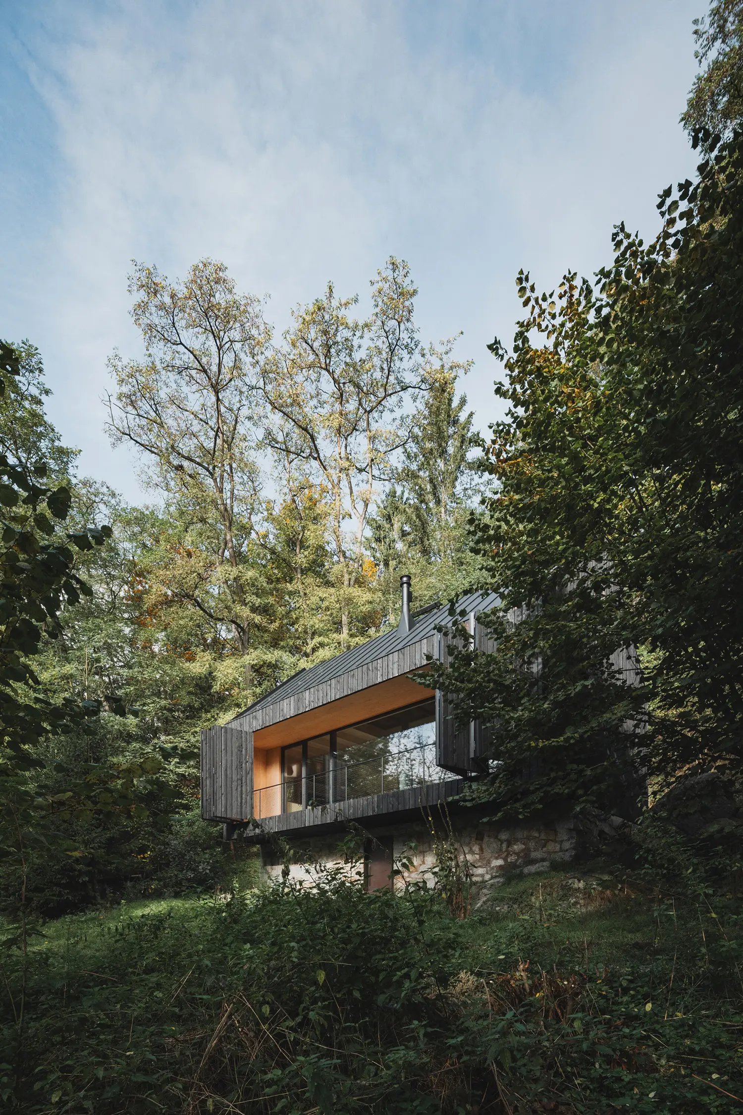 A low-angle perspective of the cabin showing the stone foundation and the large river-facing window with open shutters.