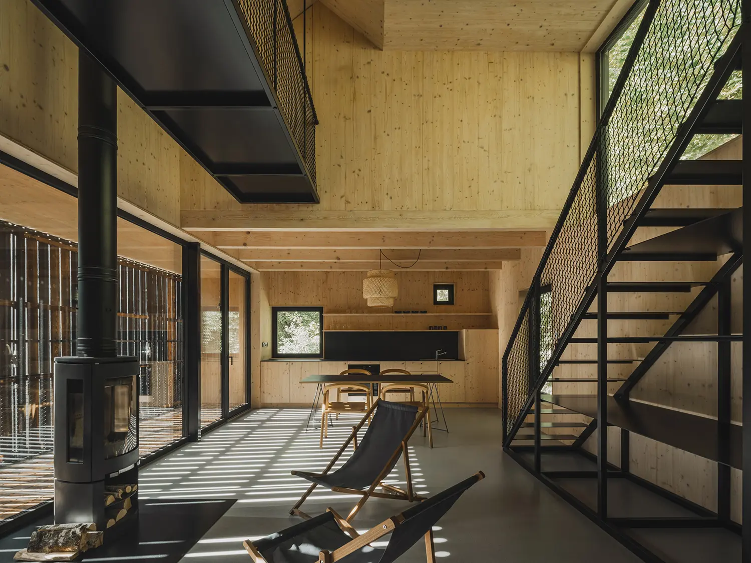 Interior of a modern wood-lined cabin featuring a black metal fireplace, staircase, and large windows with forest views.