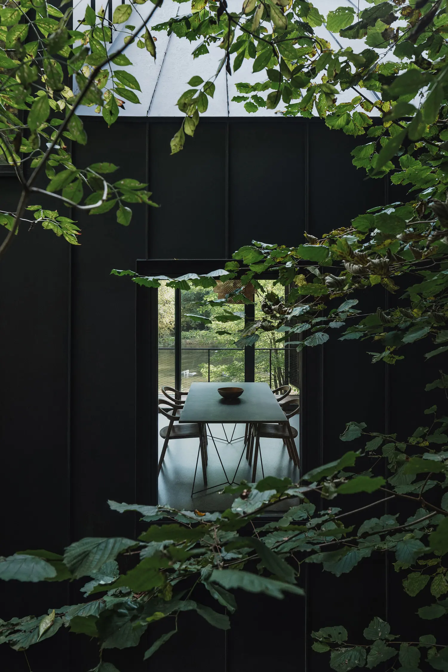 A view through a rectangular window showing a dining table and chairs inside the cabin, framed by green leaves.