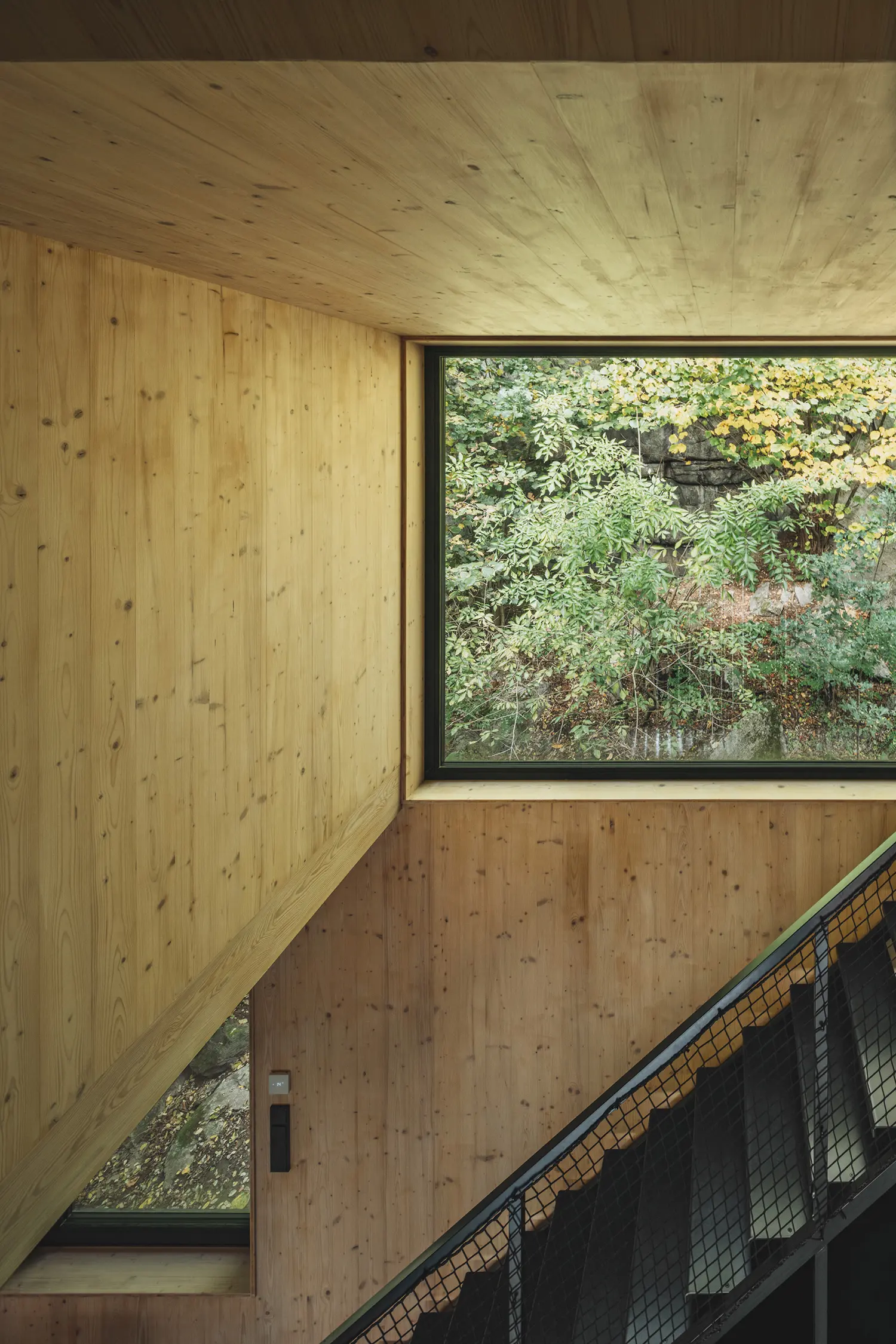 Detail of a black metal staircase against light wood walls with a window looking out at green foliage.