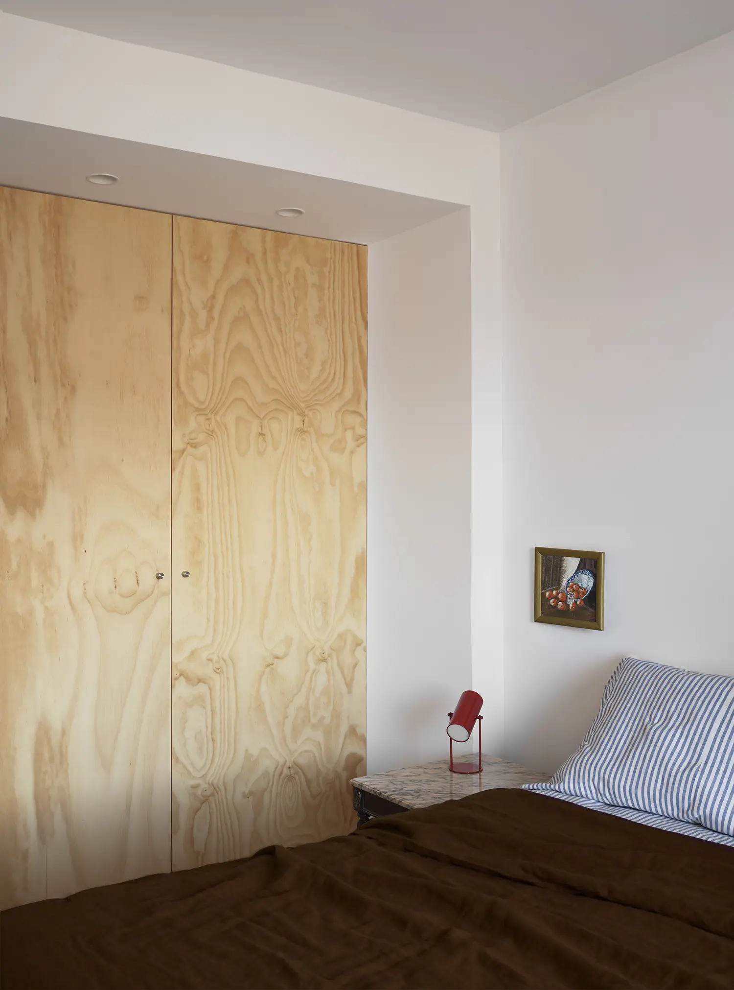 Bedroom interior featuring a plywood wardrobe, a marble nightstand, and a bed with striped pillows.