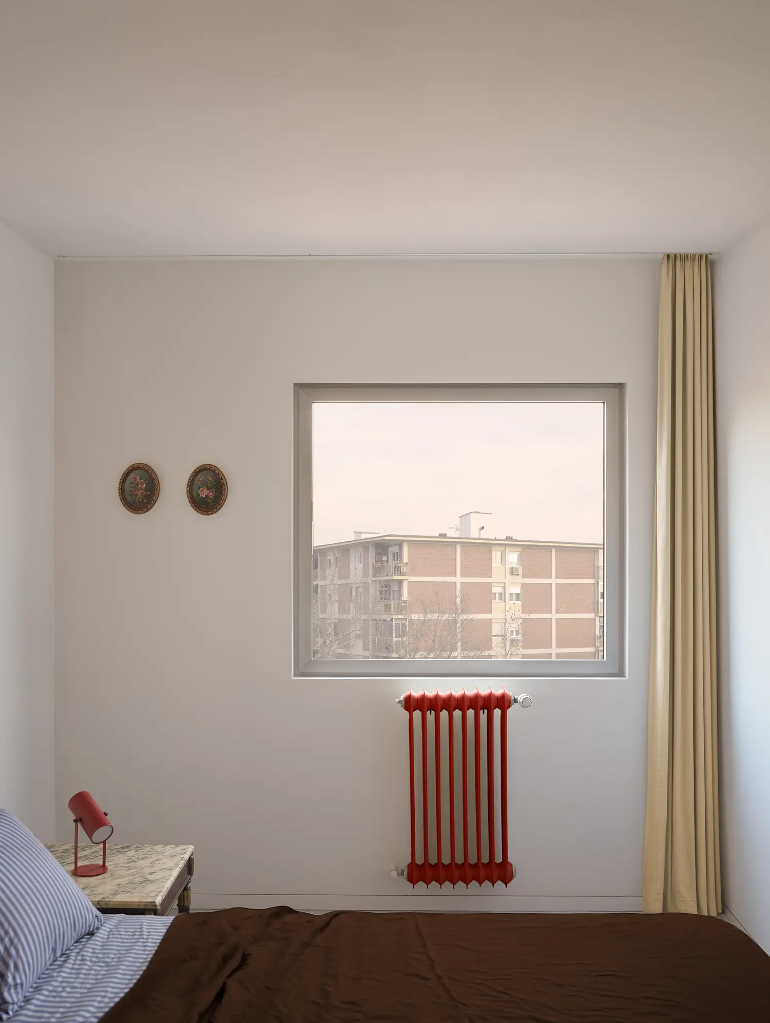 Bedroom view with a red radiator under a window looking out at a 1970s Madrid apartment block.