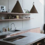 Wide angle shot of a modernist kitchen with concrete walls, two identical conical pendant lights, and a blurred human figure by the stairs.