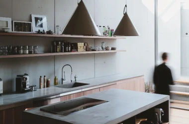 Wide angle shot of a modernist kitchen with concrete walls, two identical conical pendant lights, and a blurred human figure by the stairs.