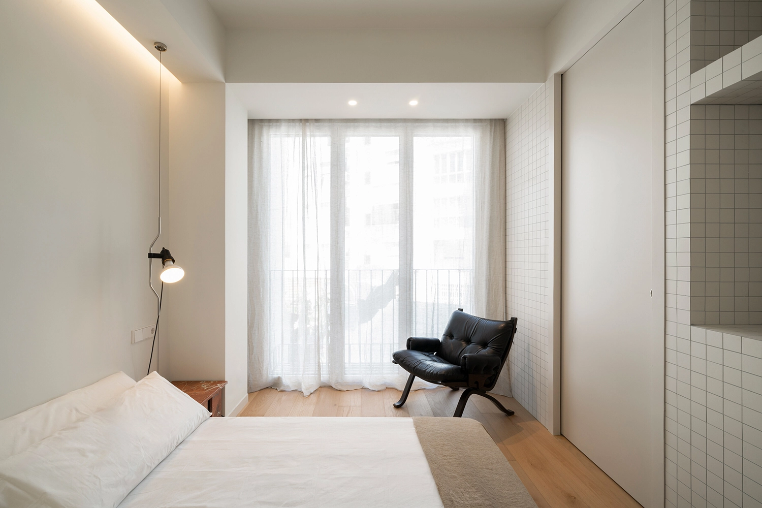 Minimalist bedroom with a white bed, black lounge chair, and a wall of white square tiles.