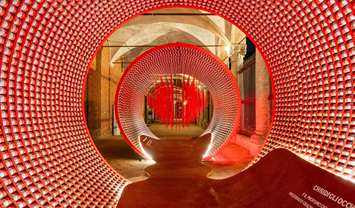 Interior view of a circular tunnel made of red tomato cans with red hanging strands in the distance at night.