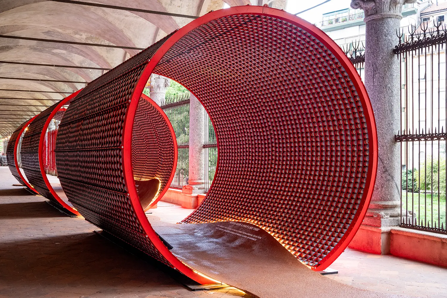 Side view of three large cylindrical structures made of red cans under a classical stone portico.