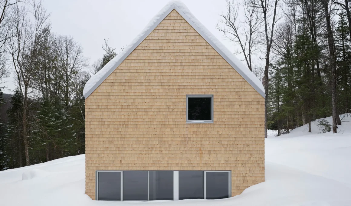Symmetrical gable end of a cedar-clad cabin partially submerged in deep snow in a Quebec forest.
