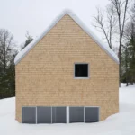 Symmetrical gable end of a cedar-clad cabin partially submerged in deep snow in a Quebec forest.