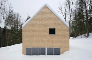 Symmetrical gable end of a cedar-clad cabin partially submerged in deep snow in a Quebec forest.