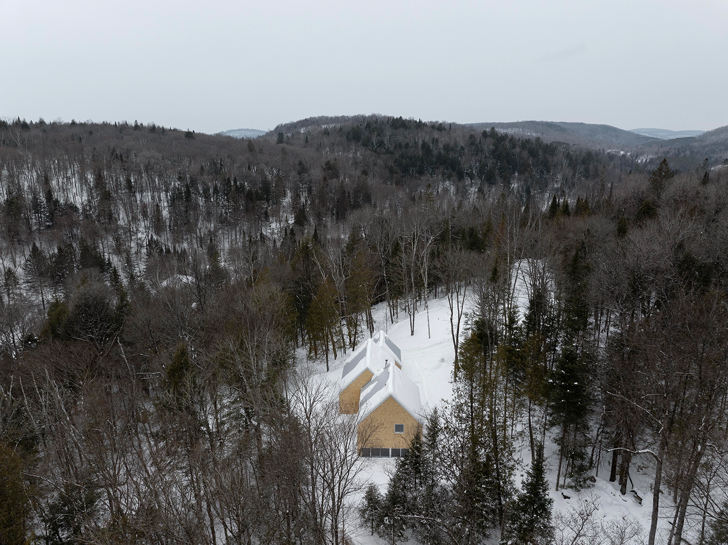 Aerial view of La Maraude cabin surrounded by a dense, snow-covered forest and rolling hills.