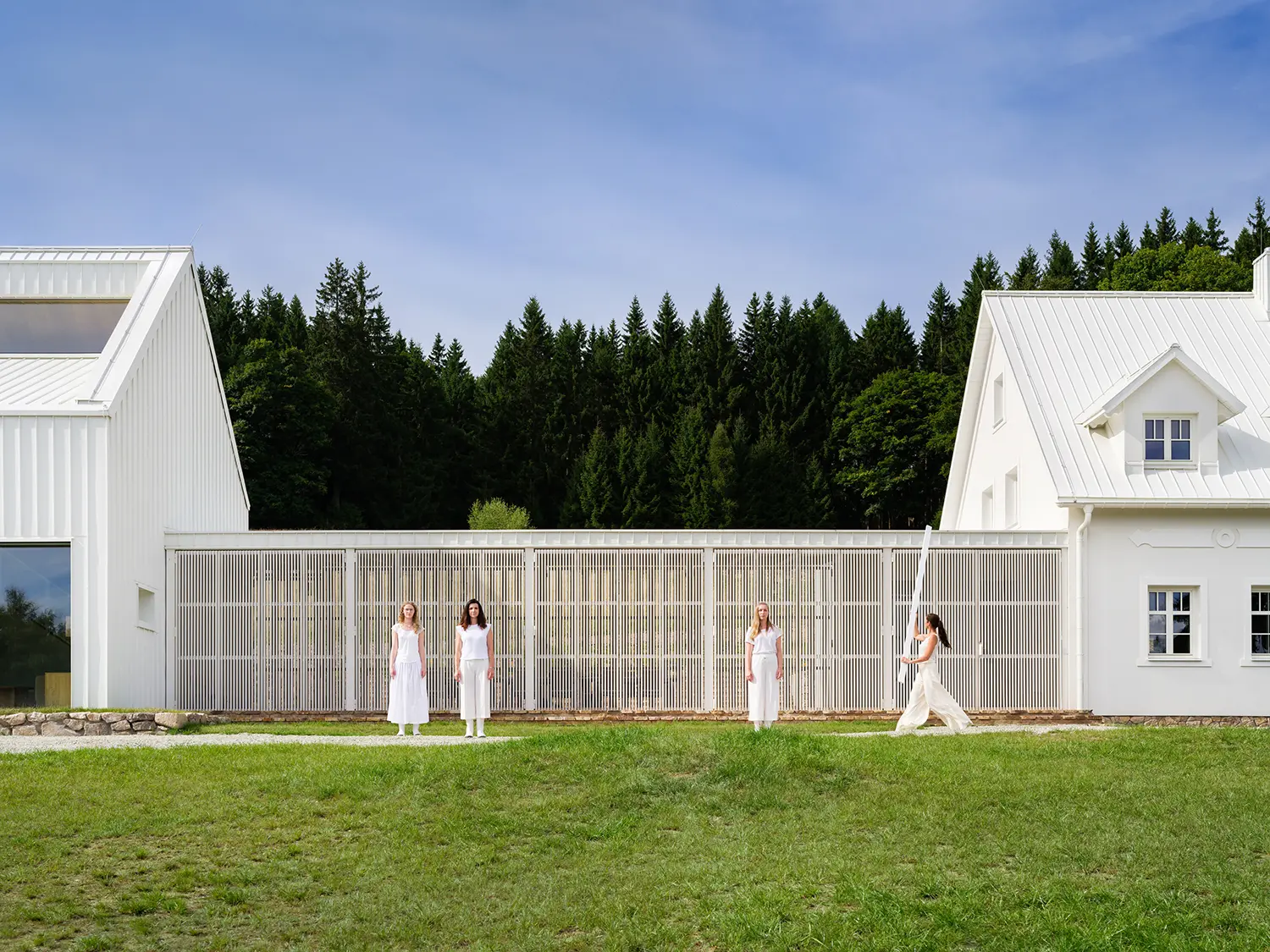 Four women in white clothing standing in front of a long slatted wooden screen connecting two white mountain buildings.