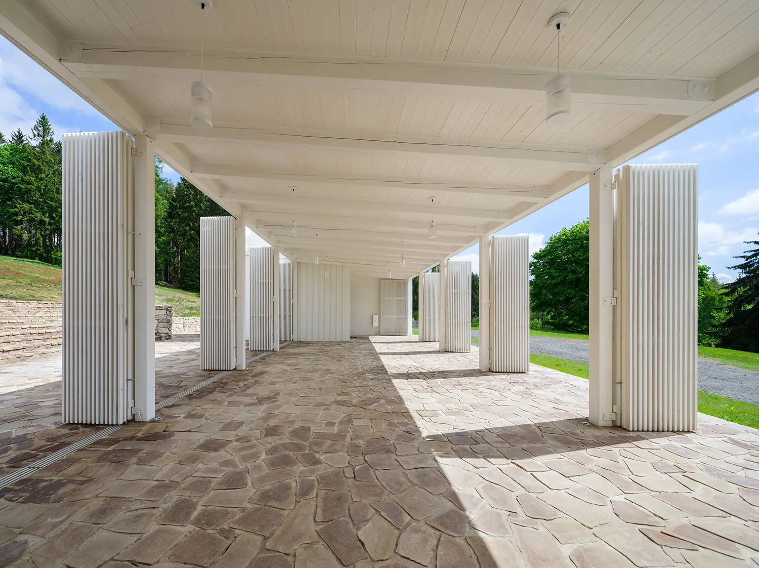 Interior view of a long covered terrace with white wooden ceilings, white pillars, and a local stone floor.