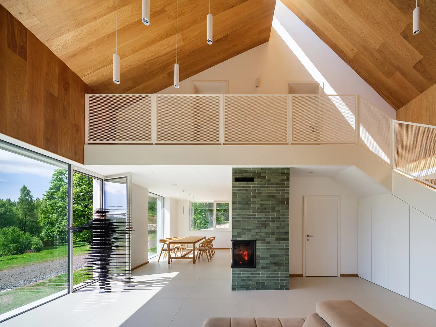 Modern double-height interior with a green tiled fireplace, wooden ceilings, and large windows overlooking the forest.