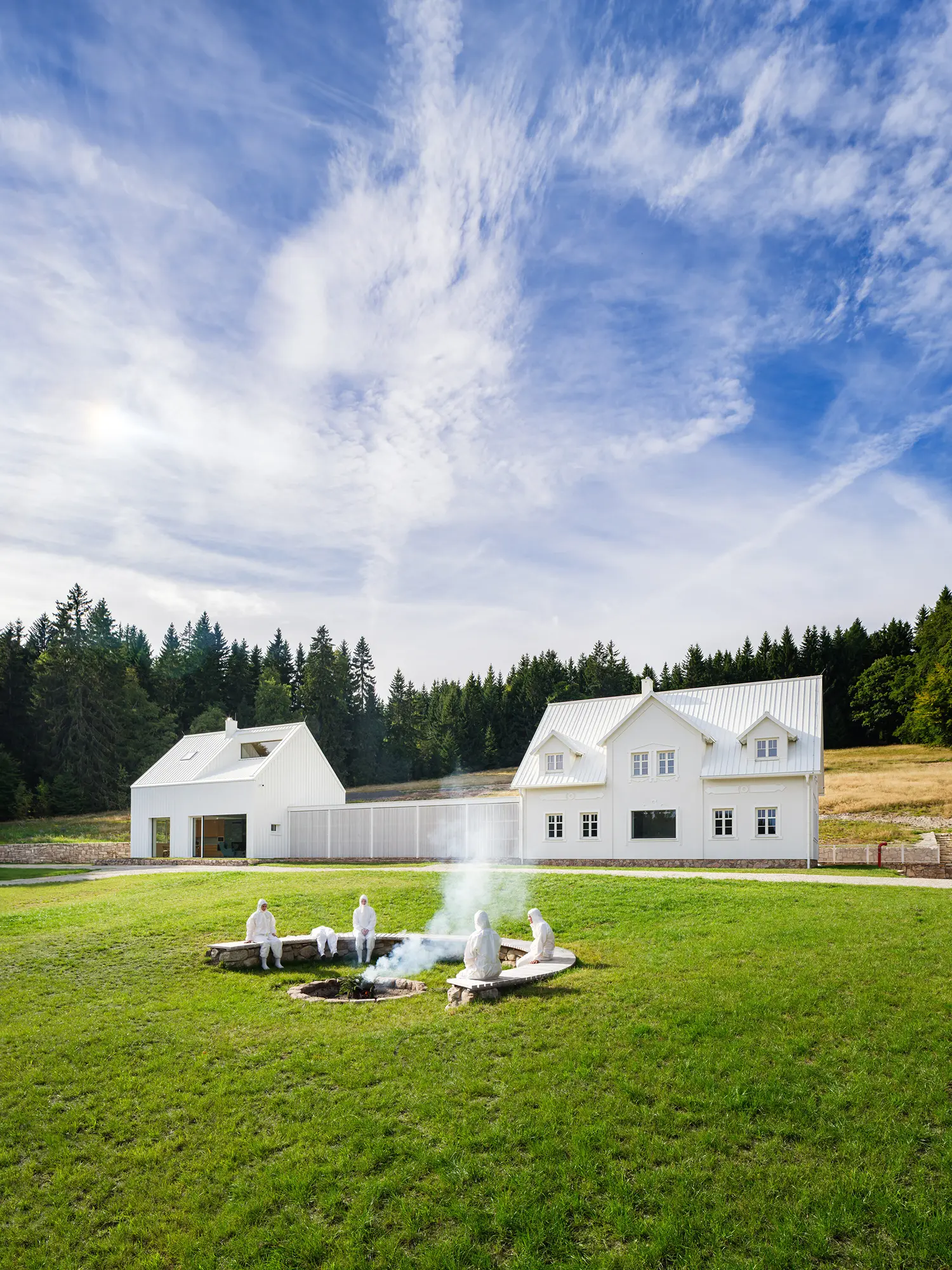 Figures in white sitting around a stone fire pit in a meadow with the white homestead and forest in the background.