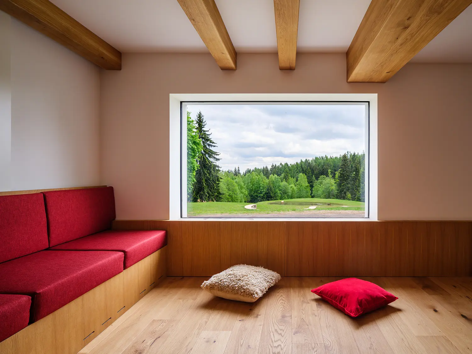Minimalist interior nook with a large square window framing a forest view, red seating, and light wood panels.