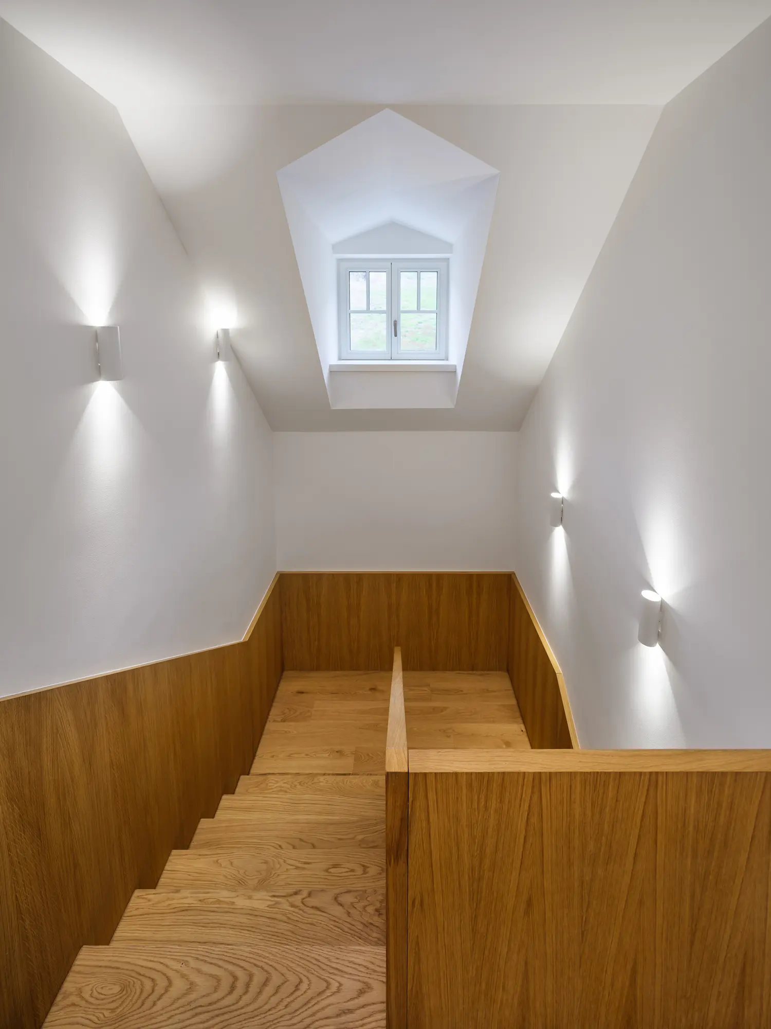 A minimalist wooden staircase leading upwards into a white gabled space with a small dormer window.