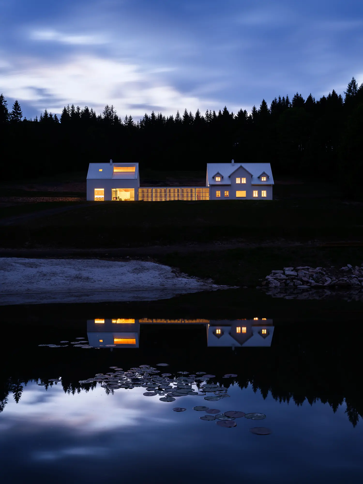 The white homestead at twilight with warm light glowing from the windows and through the slatted screen.