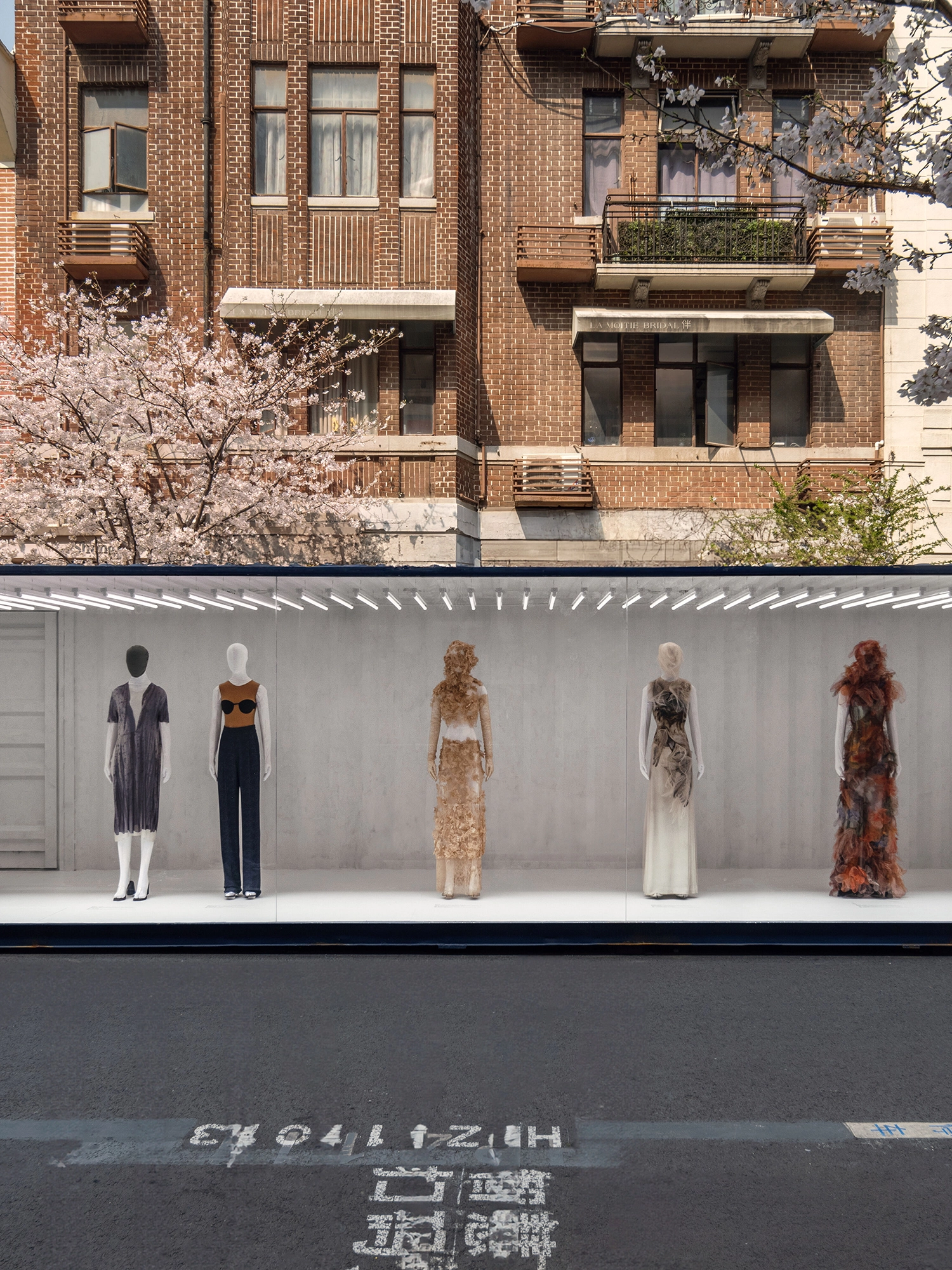 Wide shot of five mannequins in a glass-fronted shipping container positioned on a city street in front of a brick apartment building.
