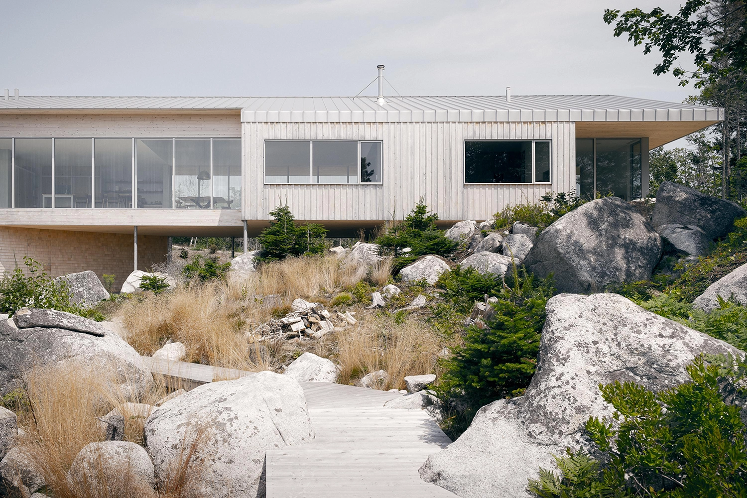 Exterior view of a long, wooden coastal home integrated into a landscape of large boulders and tall grass.