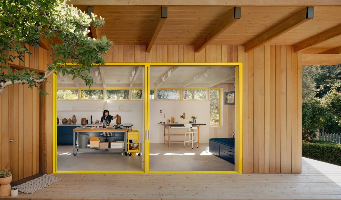 Wide sliding glass doors with yellow frames opening from a wood-clad studio onto a timber deck.