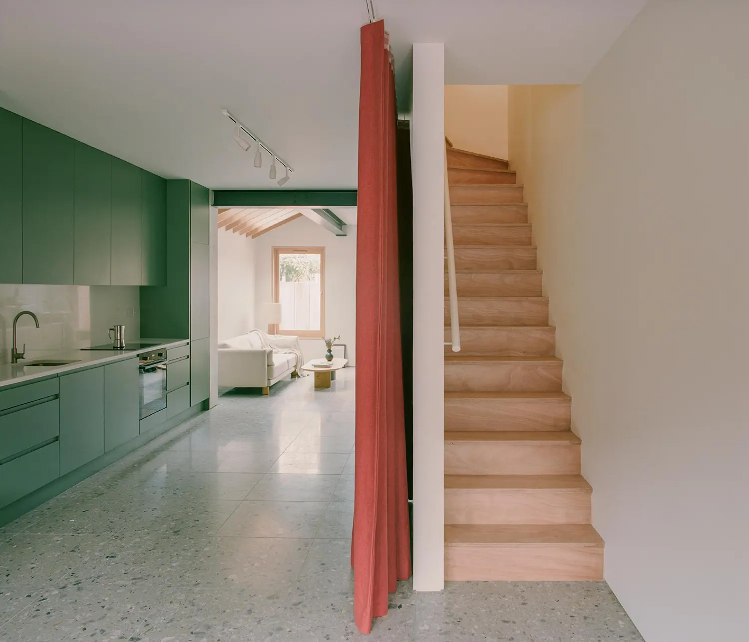Interior view showing a green minimalist kitchen, terrazzo flooring, and a red curtain separating a wooden staircase.