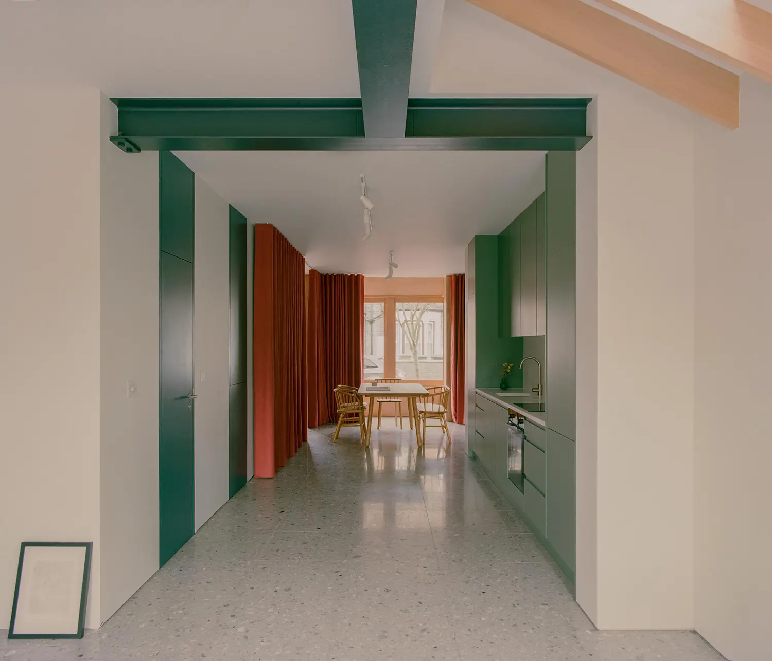 Open-plan interior looking toward a dining area with green structural beams, sage cabinets, and red curtains.
