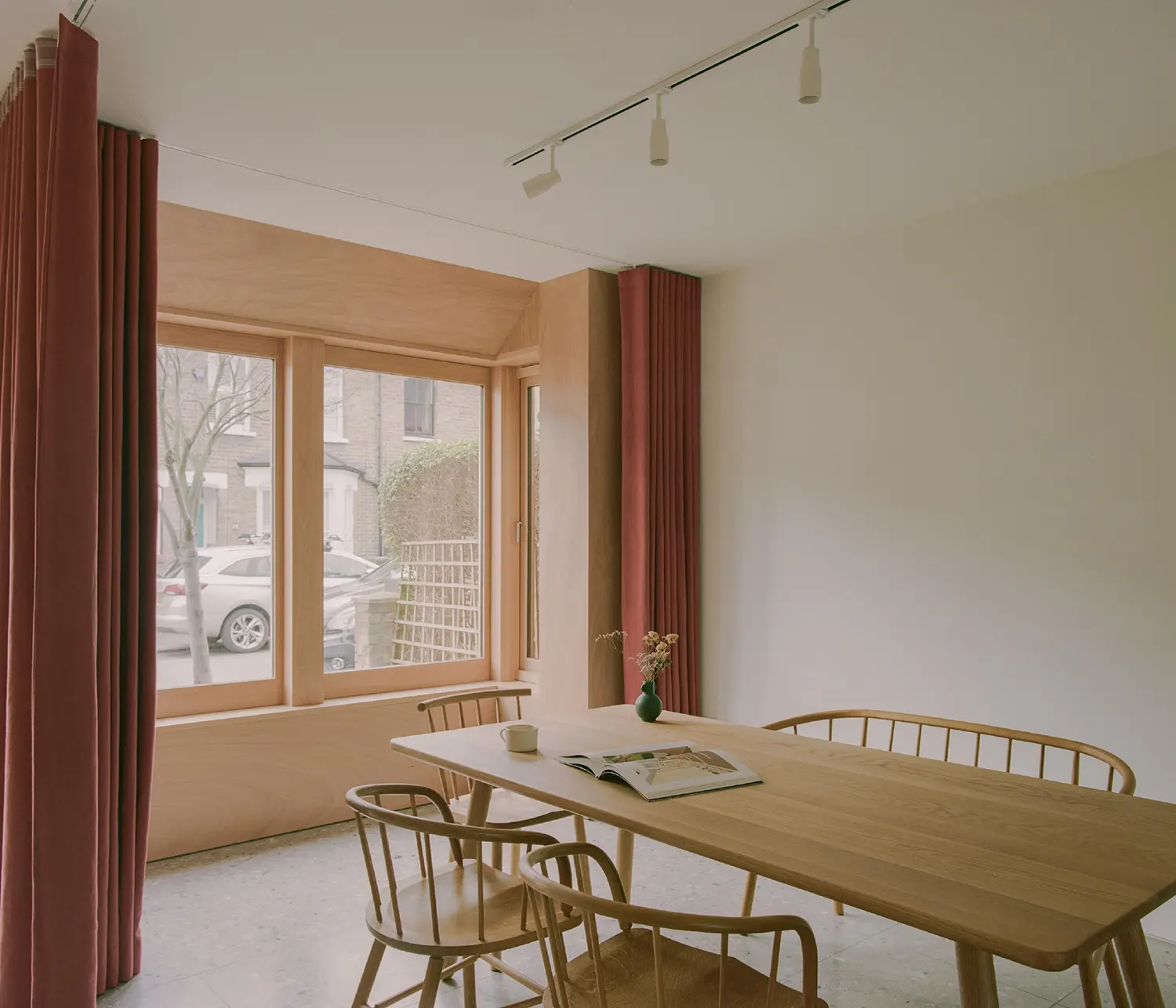Bright dining area with a light wood table, spindle chairs, and large timber-framed windows overlooking the street.