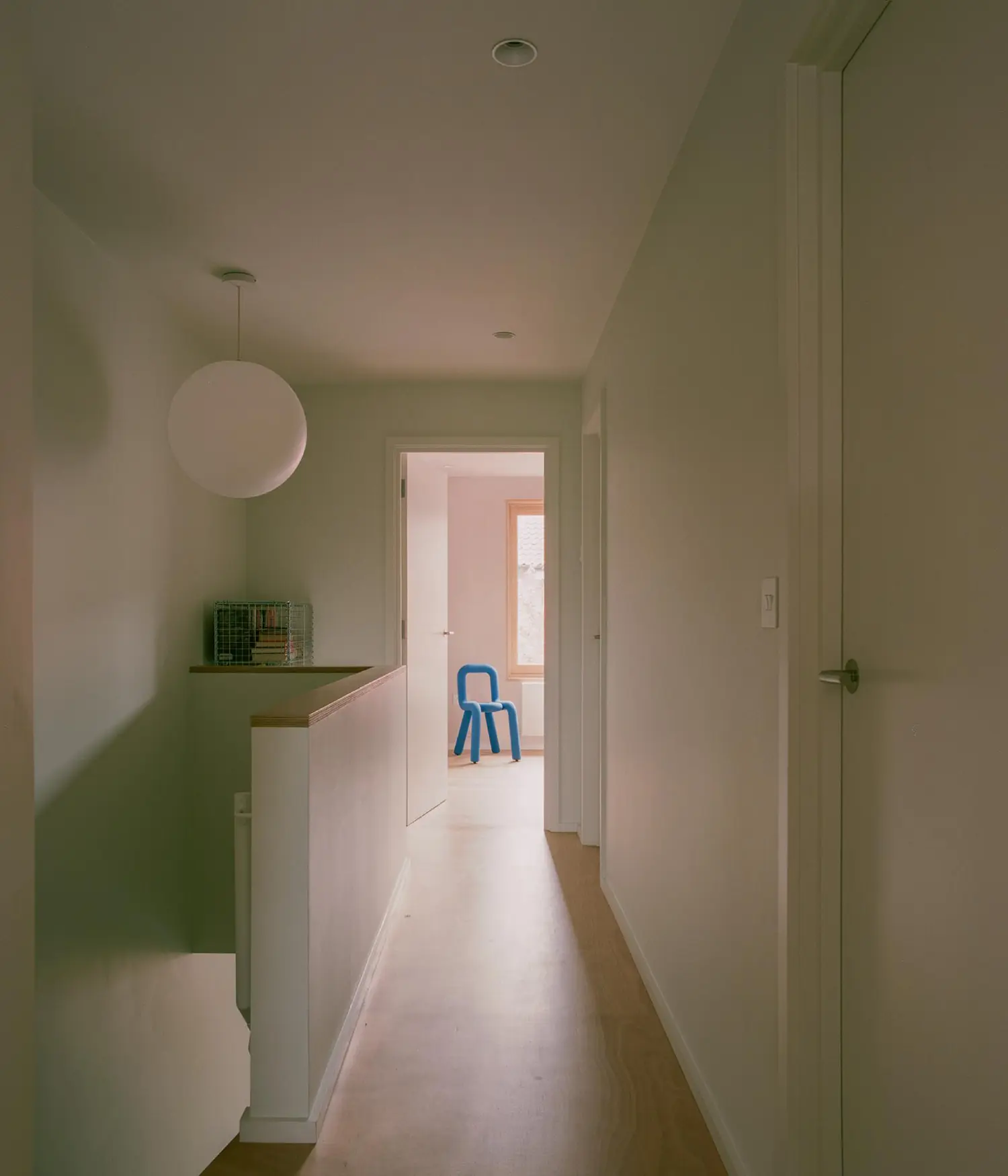 A minimalist white hallway at first-floor level featuring a round pendant light and a blue sculptural chair in the distance.