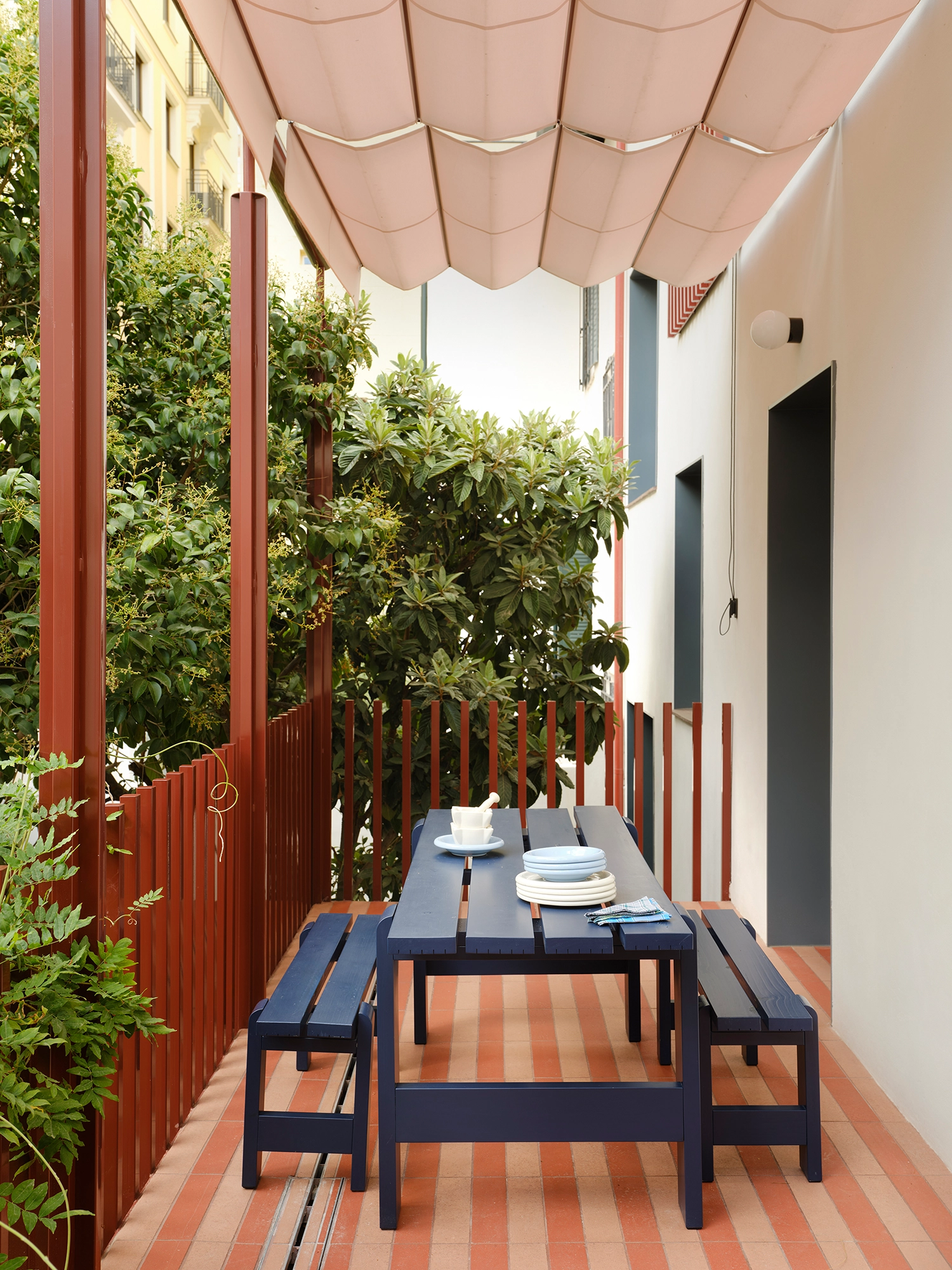Narrow outdoor terrace with a blue wooden dining table, striped terracotta floor, and red metal railings.
