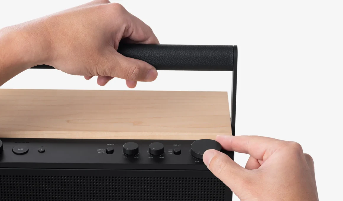 Close-up of hands interacting with the leather handle and analog volume knob of a black Fender ELIE speaker with a maple wood top.