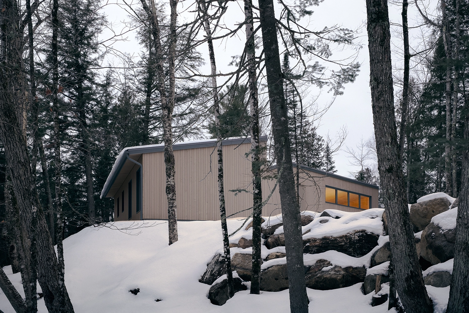 Exterior of a timber-clad house perched on a snowy escarpment among tall trees in Quebec.