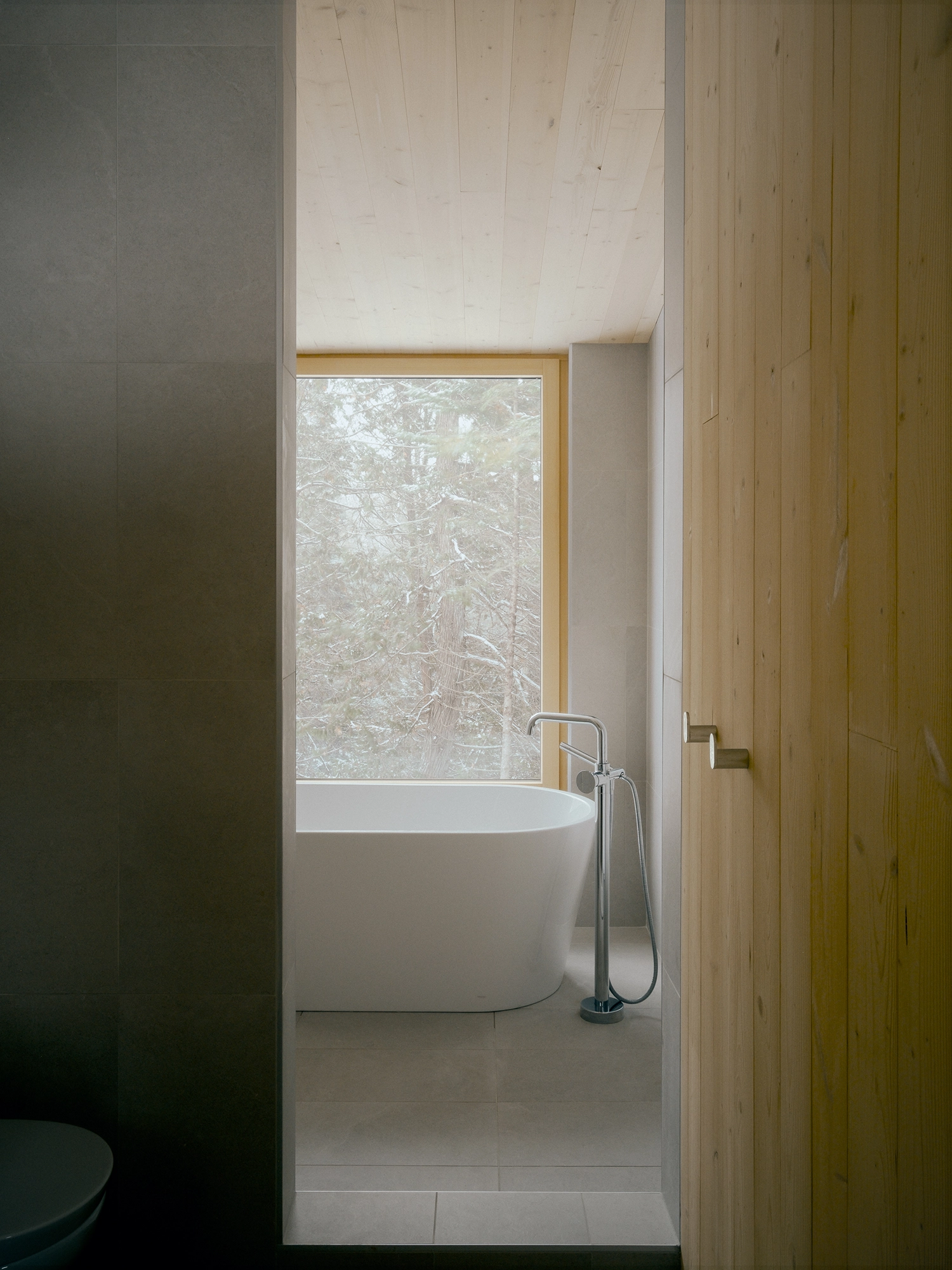 Minimalist bathroom featuring a white freestanding bathtub positioned in front of a window looking out at snowy trees.