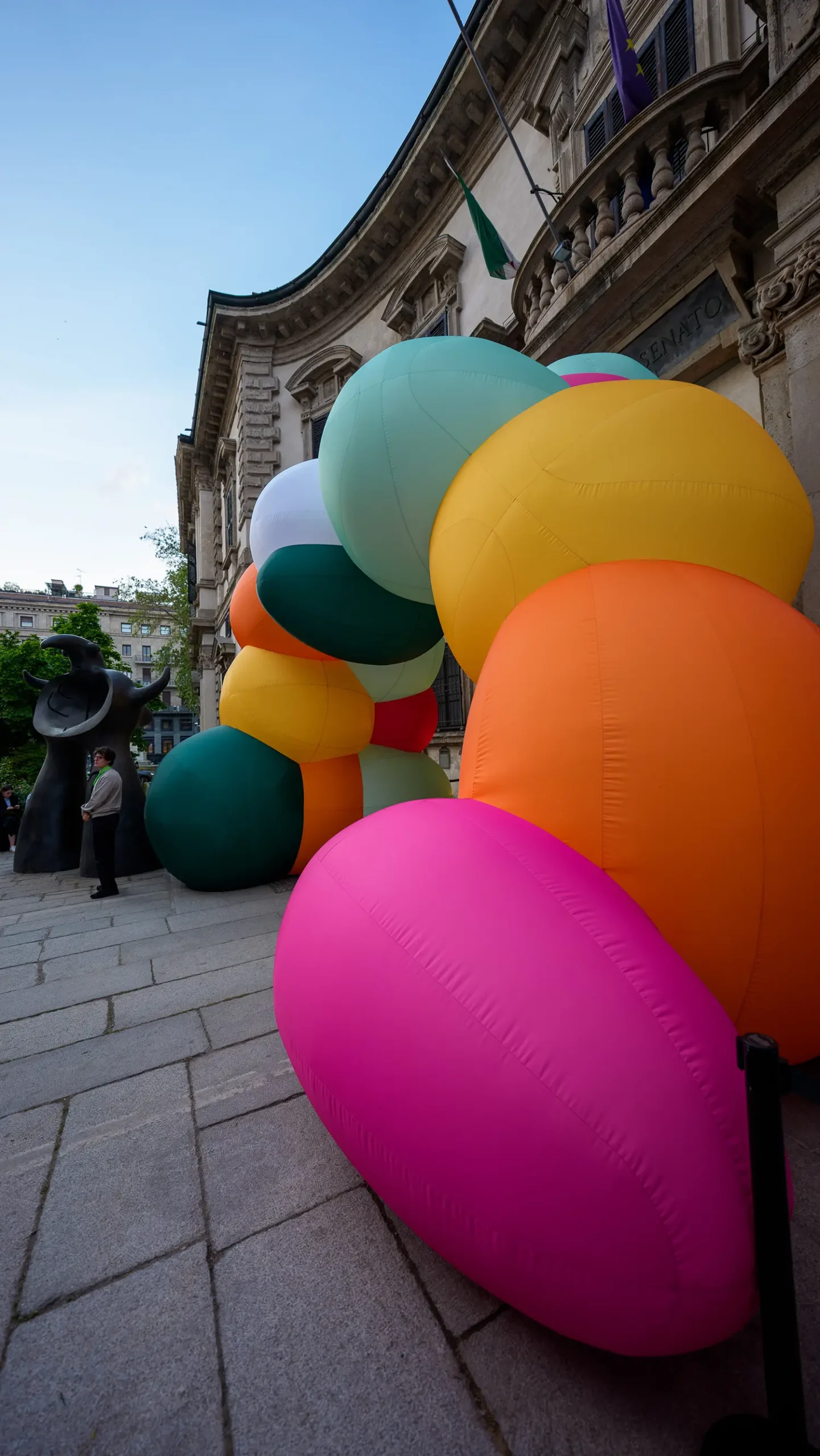 Vibrant, large-scale inflatable spheres in pink, orange, yellow, and green clustered against the stone facade of the Baroque Palazzo del Senato.