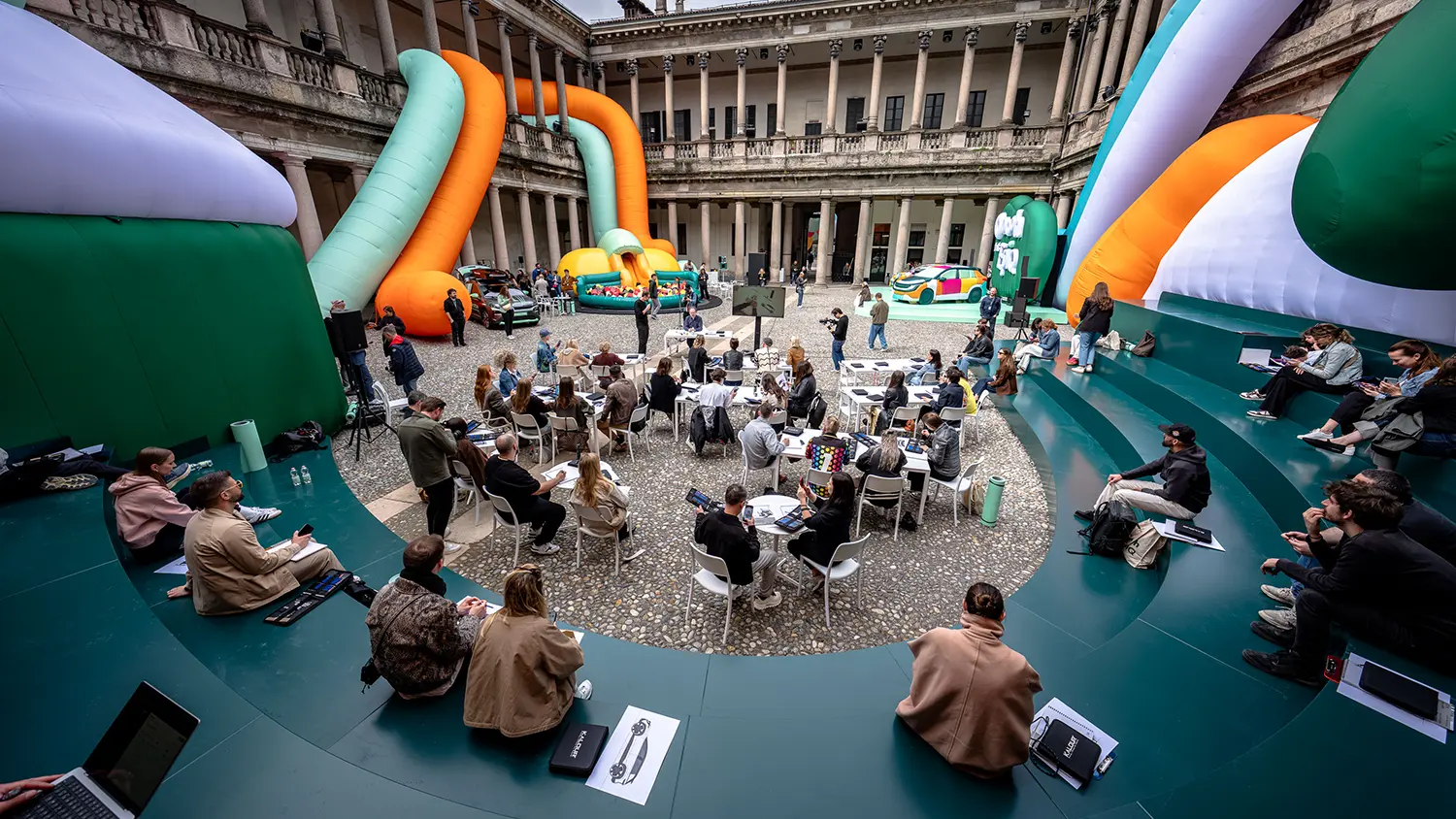 A wide shot of a lecture taking place in the Palazzo del Senato courtyard, with attendees sitting in a circular teal auditorium surrounded by orange and blue inflatable tubes.